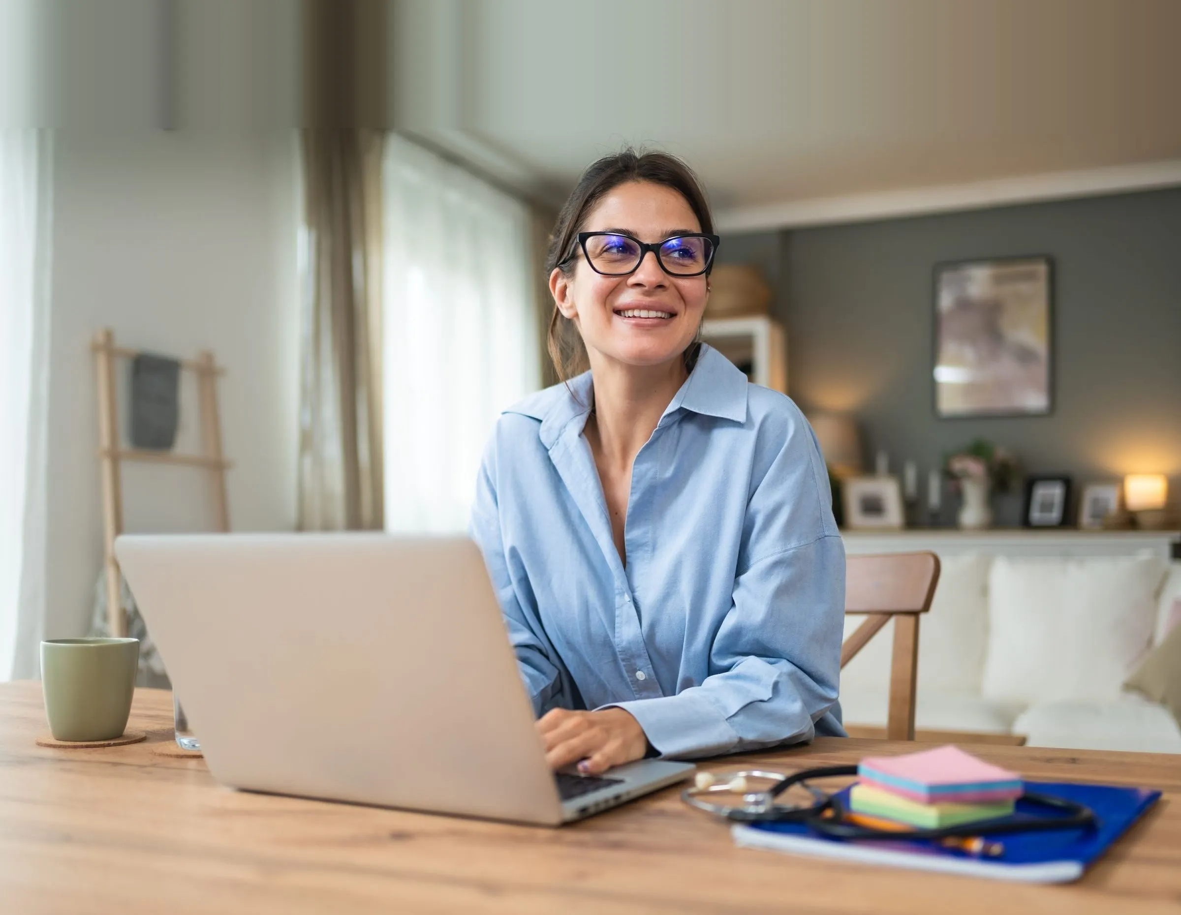 Smiling clinician in glasses working on a laptop at a home desk with a stethoscope nearby